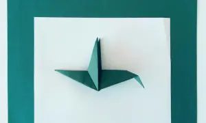 Zielona rzeźba z papieru, żuraw origami, prezentuje się na białym tle otoczonym ciemnozielonymi kartkami.