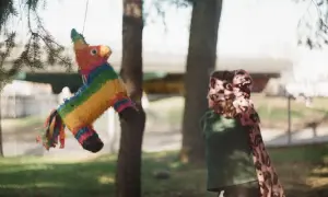Kolorowa piniata w kształcie konia wisi na drzewie. Dziecko z zawiązanymi oczami przygotowuje się do uderzenia, ucząc się, jak zrobić piniatę.
