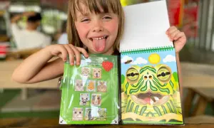 Uśmiechnięta dziewczynka pokazuje książeczkę z naklejkami zwierzątek i kolorowankę z żabą. Idealne zabawy dla 4 osób.