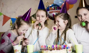 Radosne zabawy urodzinowe 12-13 lat w domu. Dziewczynki w czapkach urodzinowych dmuchają w trąbki, śmiejąc się przy torcie.