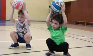 Dwóch chłopców na sali gimnastycznej, w kucki, trzymających piłki nad głowami. Radosne zabawy z piłką.