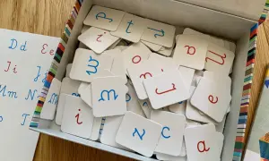 Pudełko z kartami do nauki literek przez zabawę. Na kartach litery pisane, niebieskie i czerwone. Obok tablica z literami.