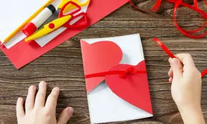 Dziecko tworzy laurkę dla mamy. Na drewnianym stole leżą nożyczki, klej, ołówek, czerwony papier i gotowa kartka z sercem przewiązana wstążką.