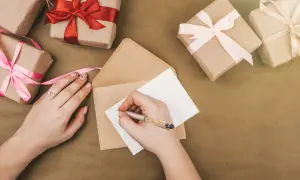 Ręce piszą zaproszenie na urodziny. Wokół leżą prezenty owinięte w papier i ozdobione wstążkami.