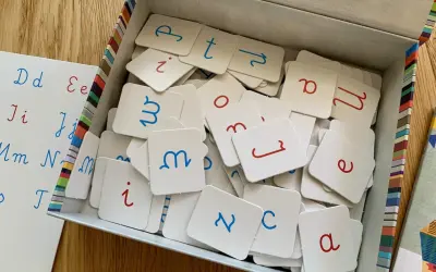 Pudełko z kartami do nauki literek przez zabawę. Na kartach litery pisane, niebieskie i czerwone. Obok tablica z literami.