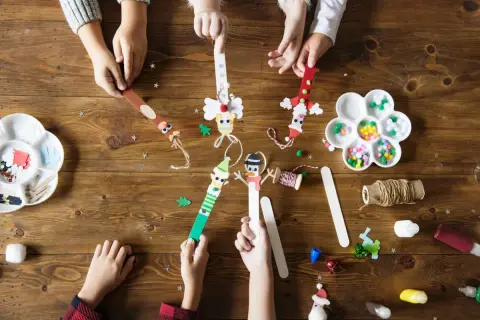 Dzieci tworzą świąteczne ozdoby z patyczków. Wesołe zabawy mikołajkowe dla dzieci na drewnianym stole.
