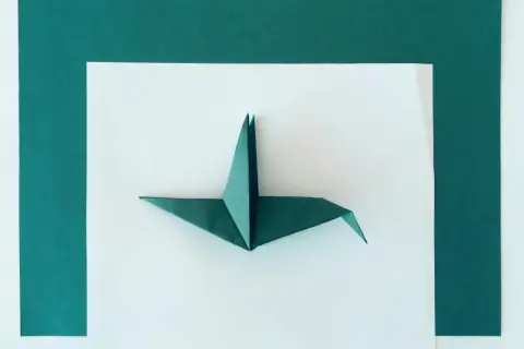 Zielona rzeźba z papieru, żuraw origami, prezentuje się na białym tle otoczonym ciemnozielonymi kartkami.
