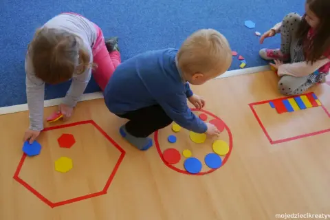 Dzieci układają kolorowe figury geometryczne w wyznaczonych obszarach.