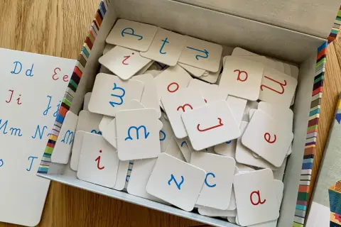 Pudełko z kartami do nauki literek przez zabawę. Na kartach litery pisane, niebieskie i czerwone. Obok tablica z literami.