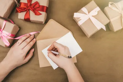 Ręce piszą zaproszenie na urodziny. Wokół leżą prezenty owinięte w papier i ozdobione wstążkami.
