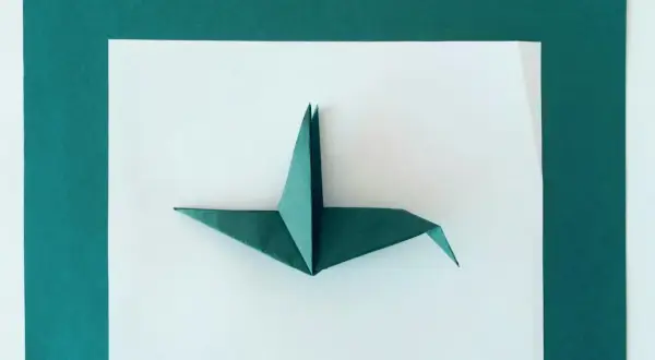 Zielona rzeźba z papieru, żuraw origami, prezentuje się na białym tle otoczonym ciemnozielonymi kartkami.