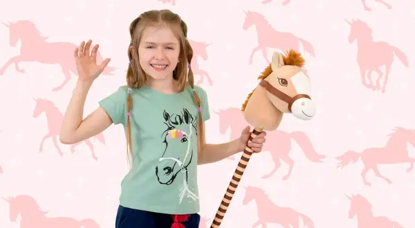 Uśmiechnięta dziewczynka z konikiem na patyku, gotowa do zabawy w hobby horsing na tle różowych jednorożców.