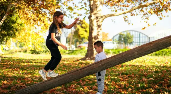 Dzieci budują tor przeszkód w ogrodzie, pokonując drewnianą kłodę. Jesienne liście dodają uroku zabawie.