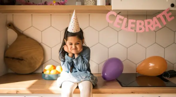 Smutna dziewczynka w czapce urodzinowej zastanawia się, co na urodziny do przedszkola zamiast słodyczy. Balony i napis "CELEBRATE" sugerują przyjęcie.