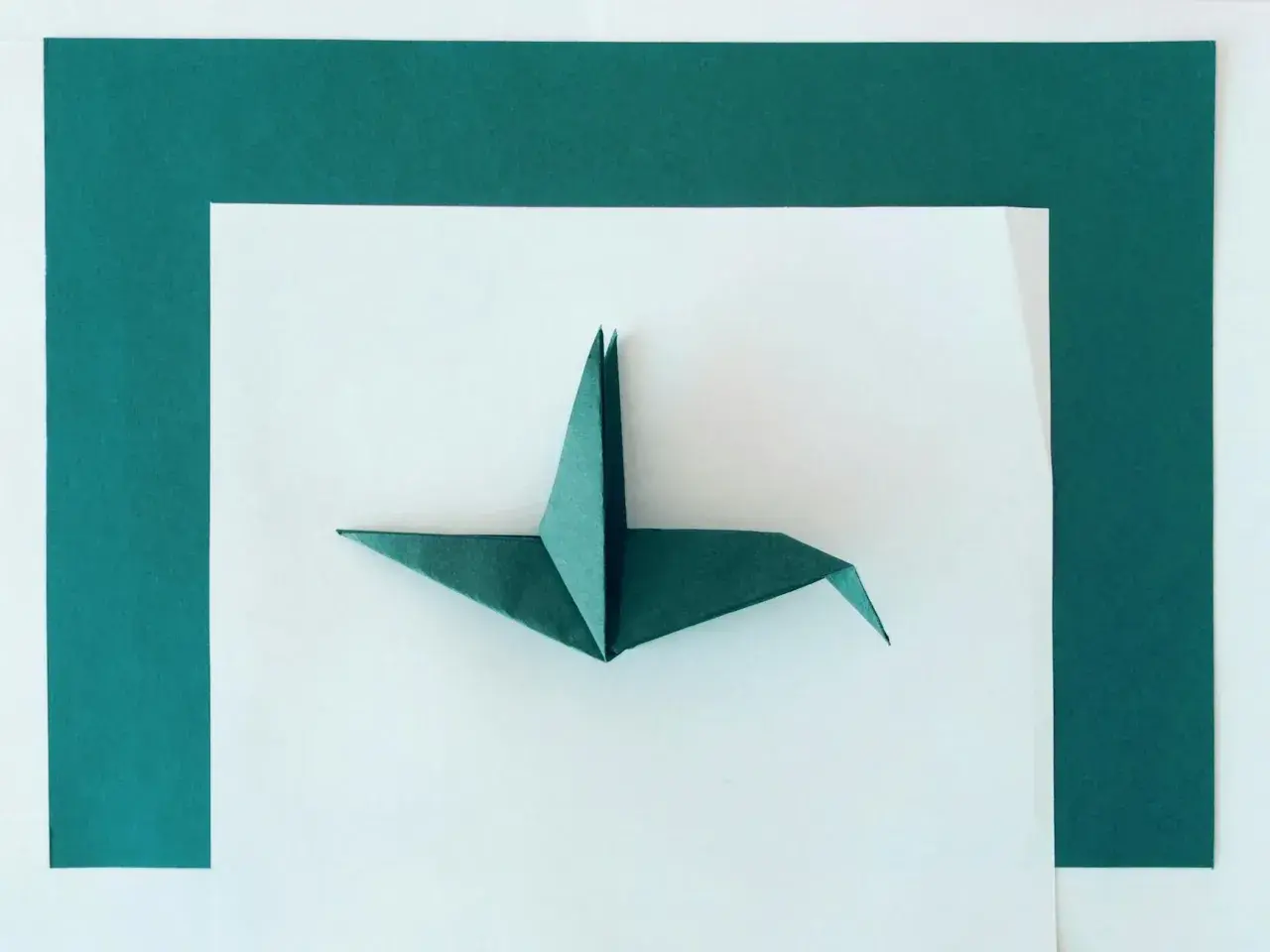 Zielona rzeźba z papieru, żuraw origami, prezentuje się na białym tle otoczonym ciemnozielonymi kartkami.
