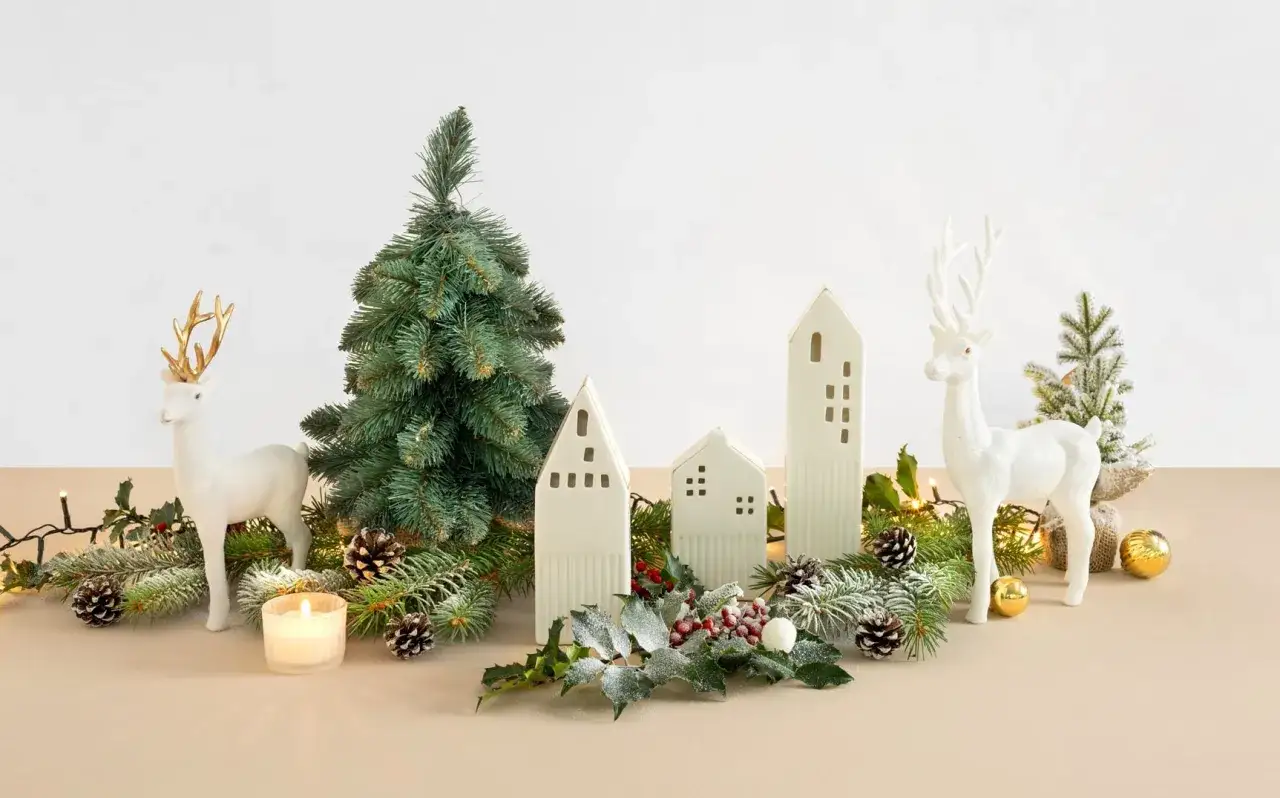 Inspiracja na stroik na st&oacute;ł wigilijny: białe renifery, choinka, ceramiczne domki, gałązki iglak&oacute;w, szyszki, świeca i lampki.