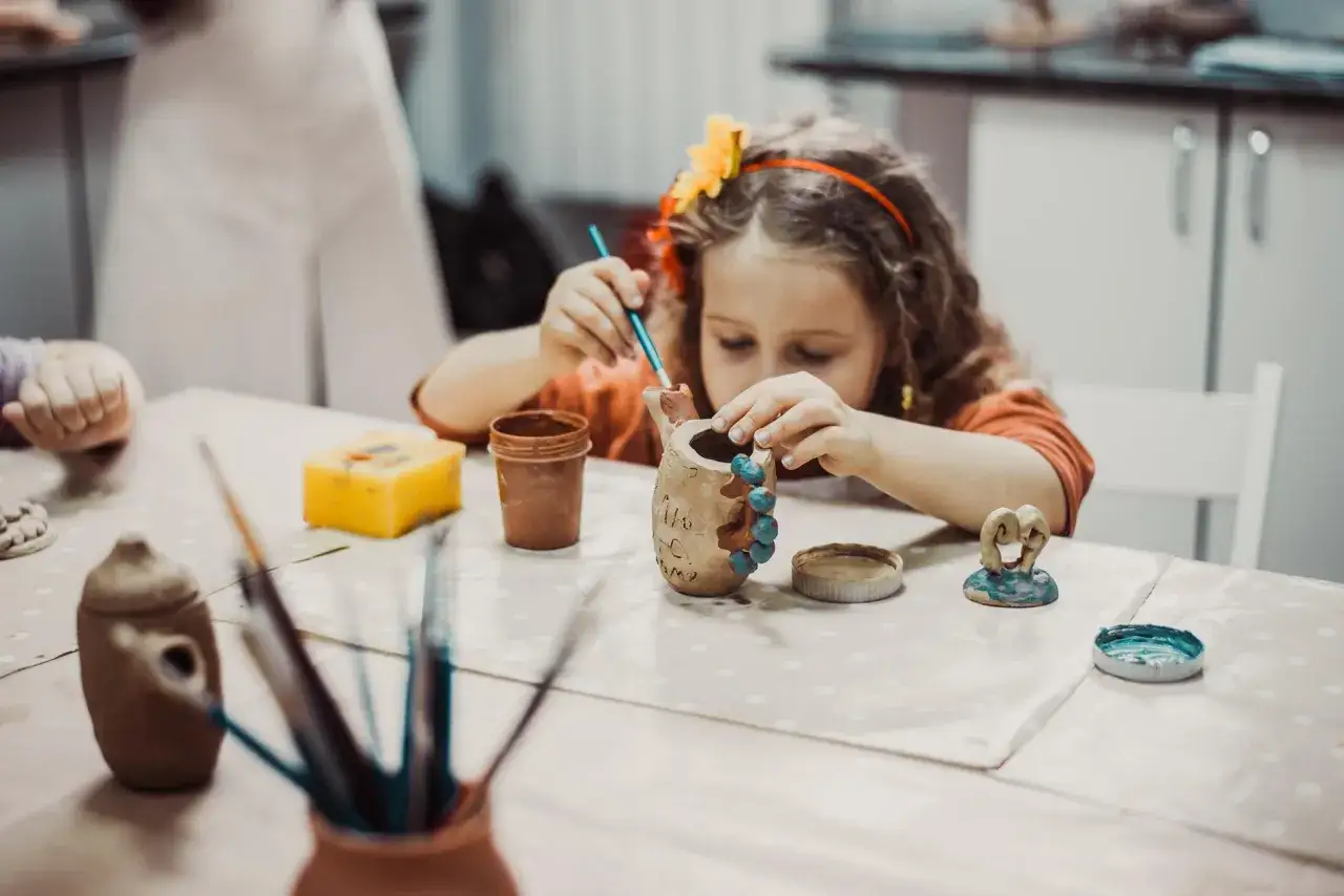 Dziewczynka z kwiatem we włosach maluje ceramiczny dzbanek. Wok&oacute;ł pędzle i inne materiały do ceramiki dla dzieci.