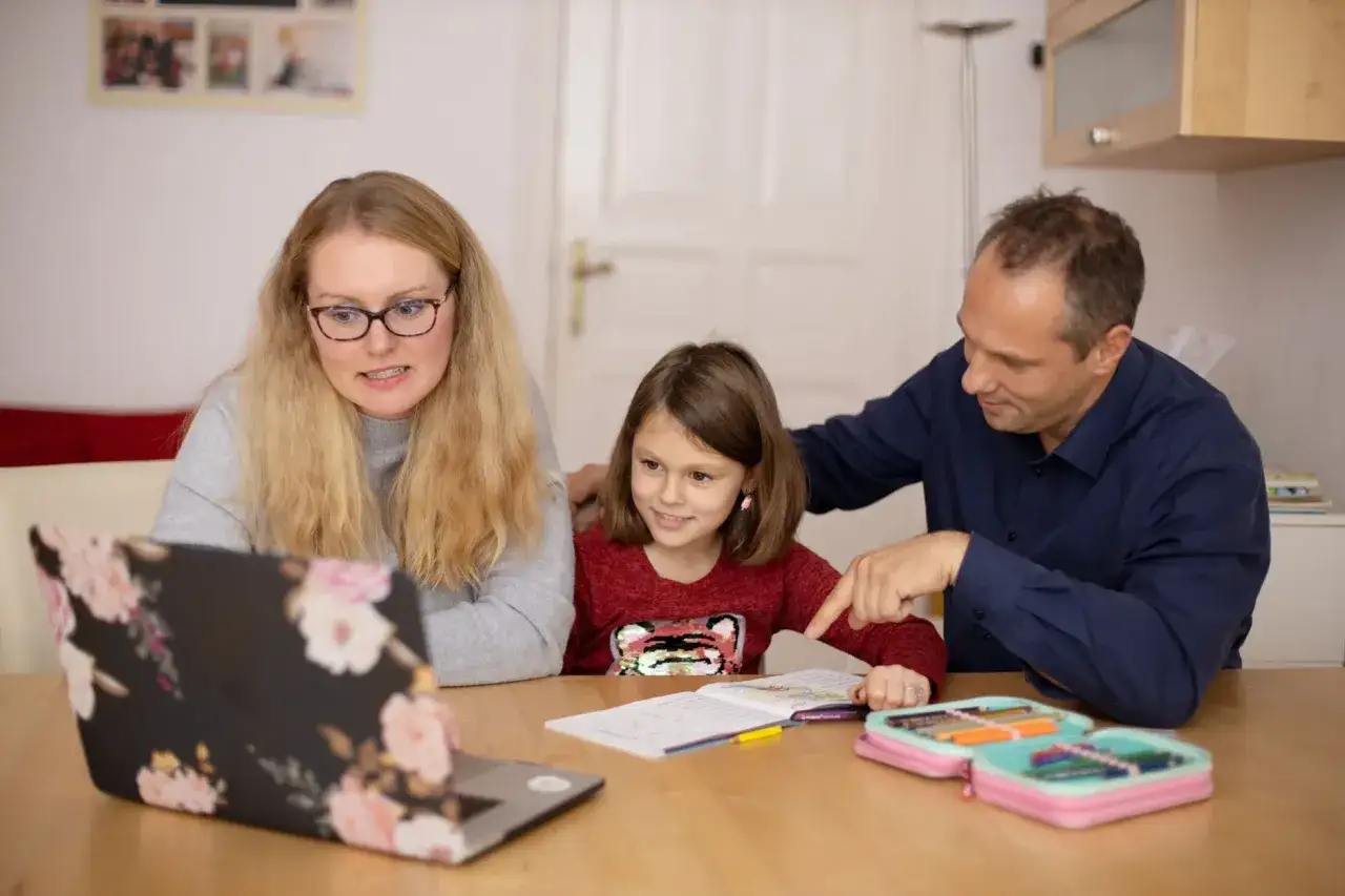 Rodzina wspólnie rozwiązuje zagadki dla dzieci, korzystając z laptopa i zeszytu. Ojciec wskazuje na zadanie, mama patrzy z uwagą.