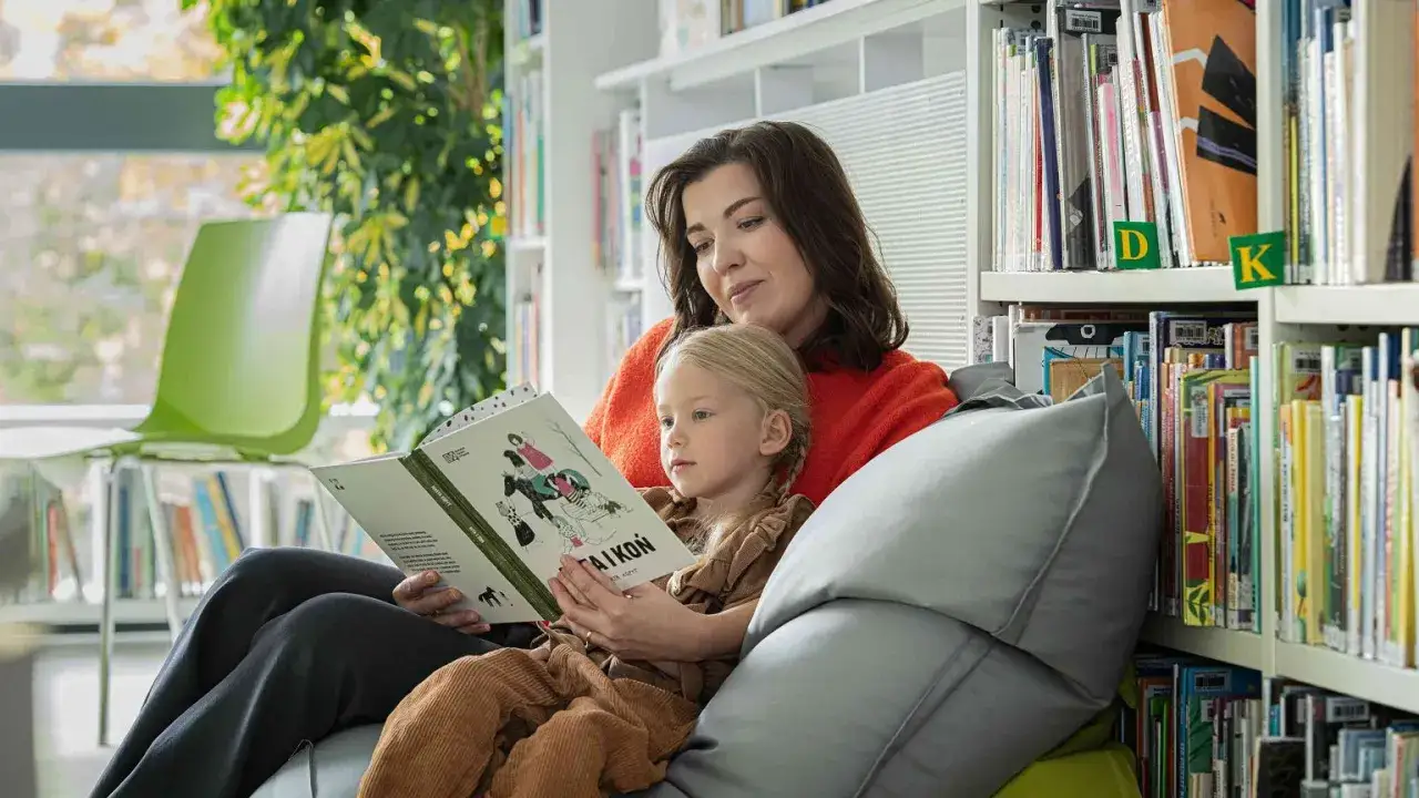 Mama czyta dziecku książkę, pokazując, jak zachecic dzieci do czytania. W tle regały pełne książek.