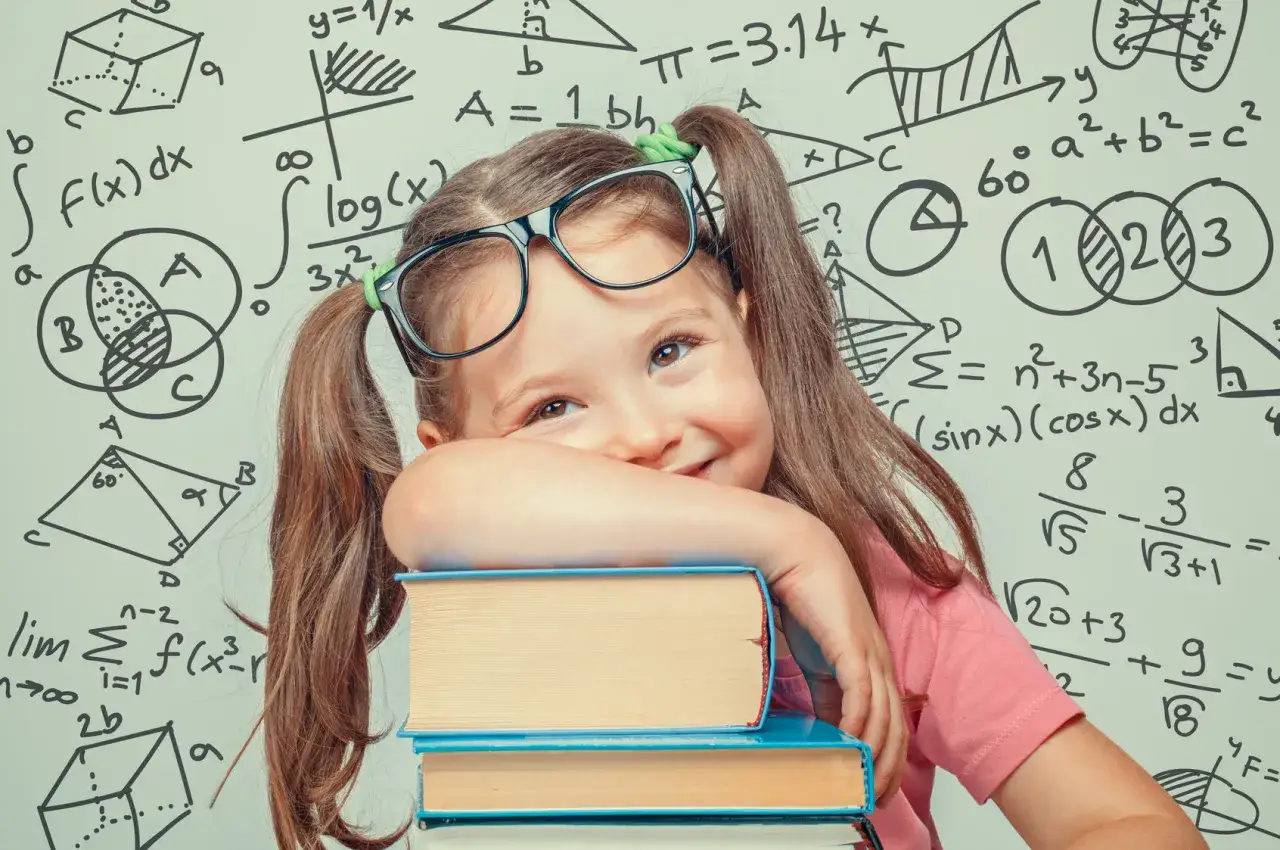 Uśmiechnięta dziewczynka w okularach opiera się o stos książek na tle tablicy z trudnymi zadaniami matematycznymi.