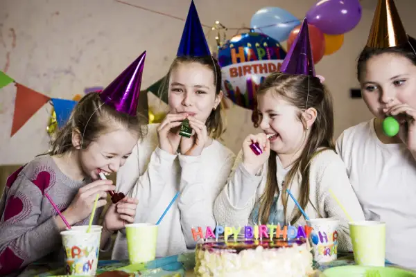 Radosne zabawy urodzinowe 12-13 lat w domu. Dziewczynki w czapkach urodzinowych dmuchają w trąbki, śmiejąc się przy torcie.