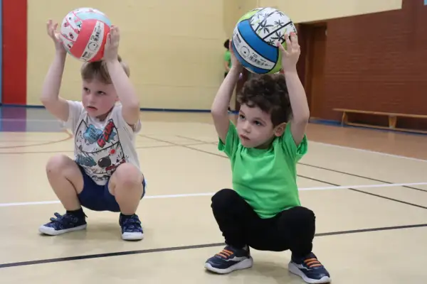Dwóch chłopców na sali gimnastycznej, w kucki, trzymających piłki nad głowami. Radosne zabawy z piłką.