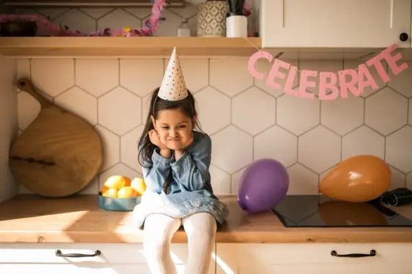 Smutna dziewczynka w czapce urodzinowej zastanawia się, co na urodziny do przedszkola zamiast słodyczy. Balony i napis "CELEBRATE" sugerują przyjęcie.